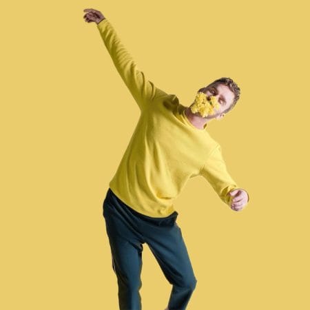 a performer with short brown hair, wearing black trousers and a long sleeved yellow top, poses against a yellow background, in their mouth is a bunch of yellow dandelions, their eyes are closed Luke Brown
