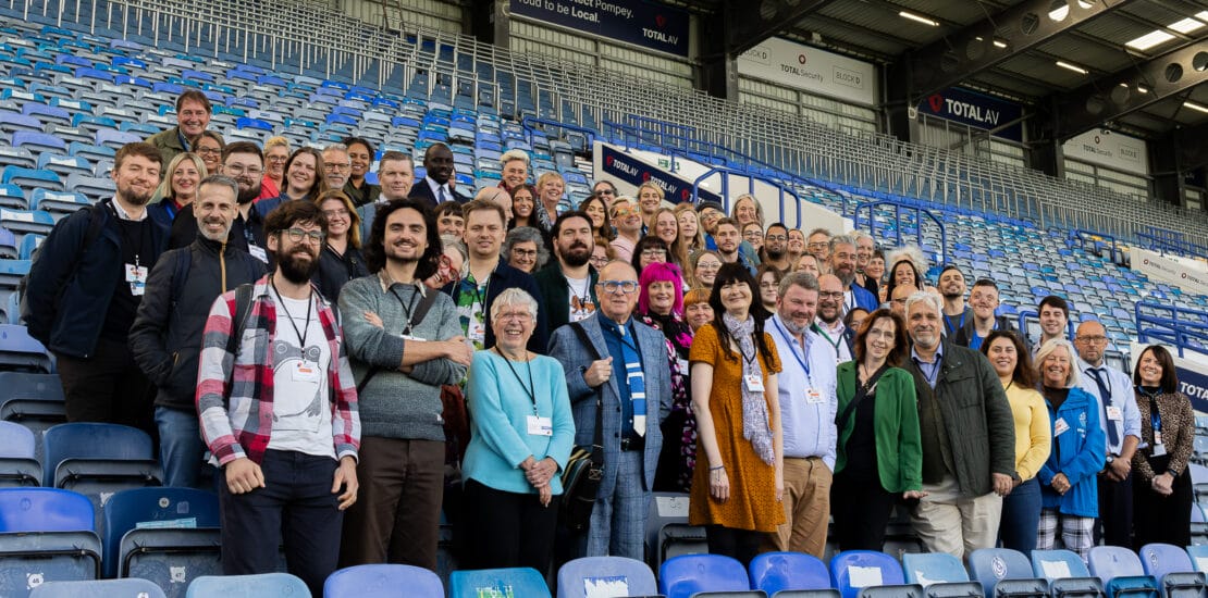a large diverse group of people pose for the camera standing in the seating area of Portsmouth football club Portsmouth Creates | We Connect Conference | 2024 allies networks and organisations