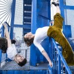 Two performers create a striking outdoor scene: one, a wheelchair user, balances on metal steps beneath a fence. The other hangs from the step by one hand, defies gravity in a moment of strength. Together, they make a dynamic contrast.