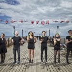 six people engaged in juggling activities on a scenic pier decorated with British Union Jack flags up behind them against a blue sky and seascape