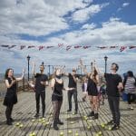 Performers dressed in black stand on a pier decorated with British flags, reaching up towards the sky while surrounded by scattered yellow juggling balls.