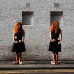 Two red-haired women in black dresses standing barefoot against a white brick wall, each holding juggling props in their hands.