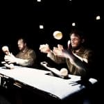 Two performers juggle props while sitting at a desk.