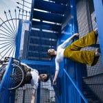Two performers create a striking outdoor scene: one, a wheelchair user, balances on metal steps beneath a fence. The other hangs from the step by one hand, defies gravity in a moment of strength. Together, they make a dynamic contrast.