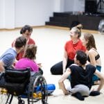 Six international performers including one wheelchair user sit indoors, on the floor in a circle listening to one of the performers, they all laugh happily
