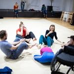 five international performers including one wheelchair user sit indoors, on the floor in a circle laughing happily with other magical memorable moving