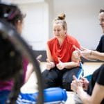 A performer in a red top and black tracksuit bottoms with brown hair tied up in a bun, puts both their thumbs up, three others do the same, they are not fully in the picture
