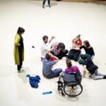 seven performers sit on the floor in a rehearsal room, they are all laughing and some are falling back or forward as they laugh, another person stands nearby watching, in black trousers and a long green cardigan, with dark mid length hair