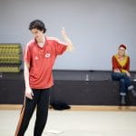 one performer in an indoor rehearsal room, in a red t-shirt and black track suit bottoms makes a dance movement while a person, slightly out of focus watches from behind