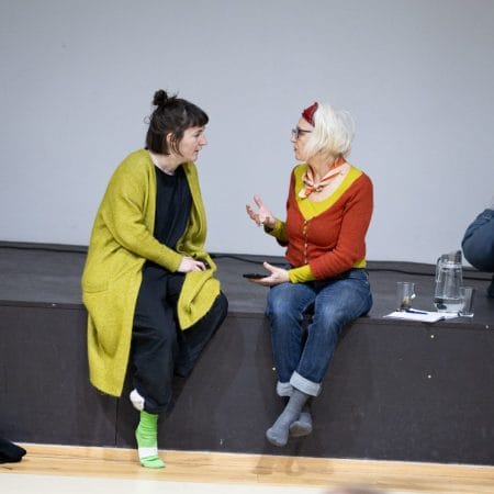 two people sit on the front of an indoor rehearsal stage, both are in socked feet and one wears blue jeans and a red cardigan, the other has black trousers and a long green cardigan, one has dark mid length hair and the other mid length blonde hair, they are talking intently to each other