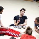 four performers sit on the floor in a rehearsal room, they are all smiling