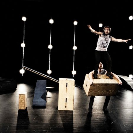 Two performers are on stage, one holding a wooden box while the other stands on it.