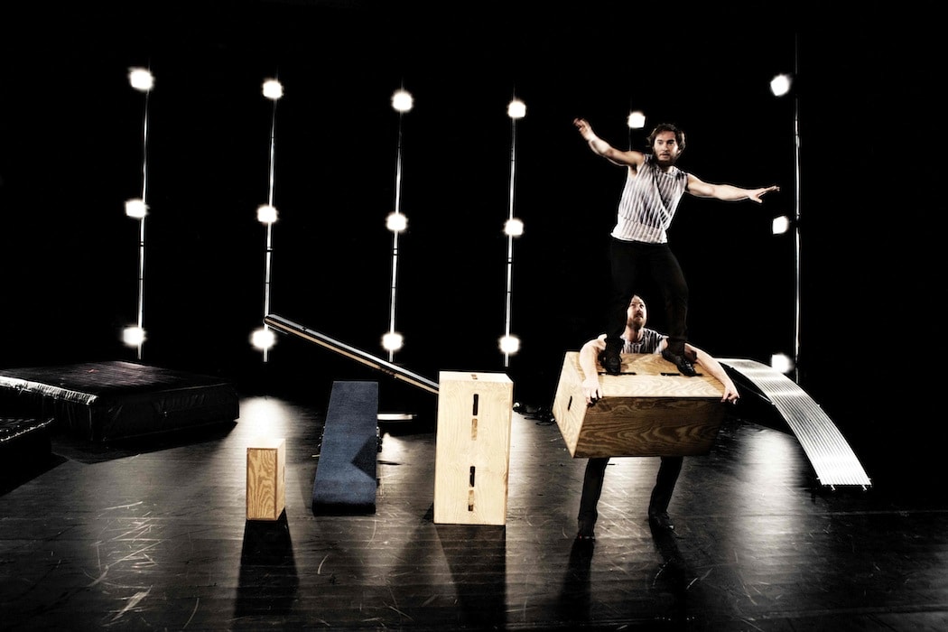 Two performers are on stage, one holding a wooden box while the other stands on it.