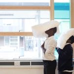 Two school children stand by a classroom window, each with a pillow balanced on their head, which hides their faces.