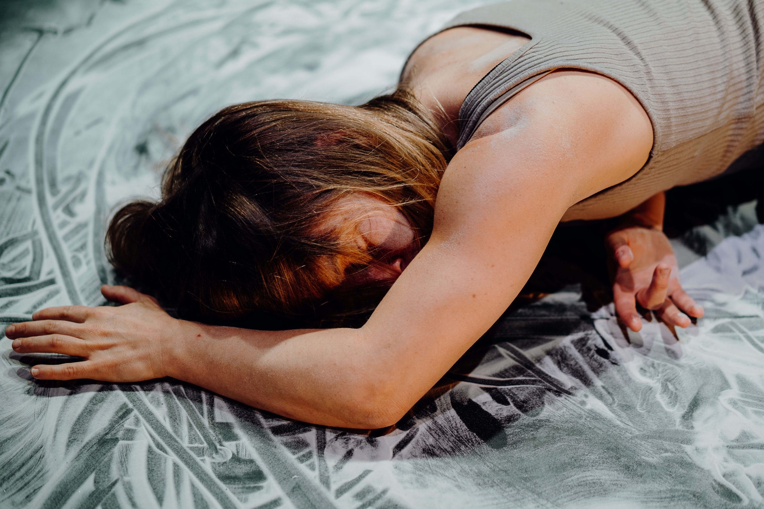 a performer in a grey vest, with flowing blonde hair is lying face down, with one arm underneath them and the other outstretched on the ground in front, the floor is covered in fake snow with marks where the performer has moved their hands