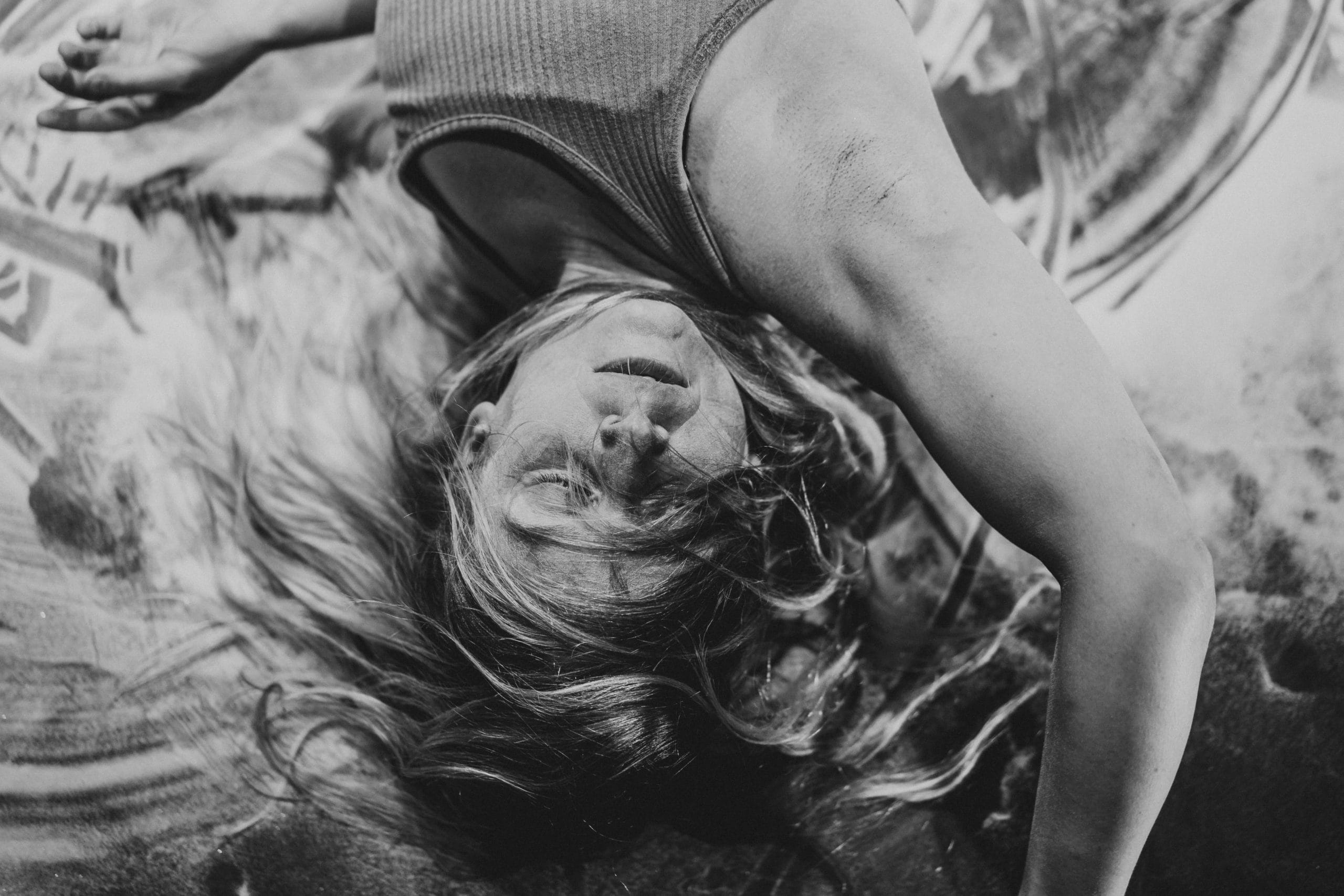 a performer in a grey vest, with flowing blonde hair is lying face up, with their eyes closed and arms outstretched on the ground, the floor is covered in fake snow with marks where the performer has moved their hands