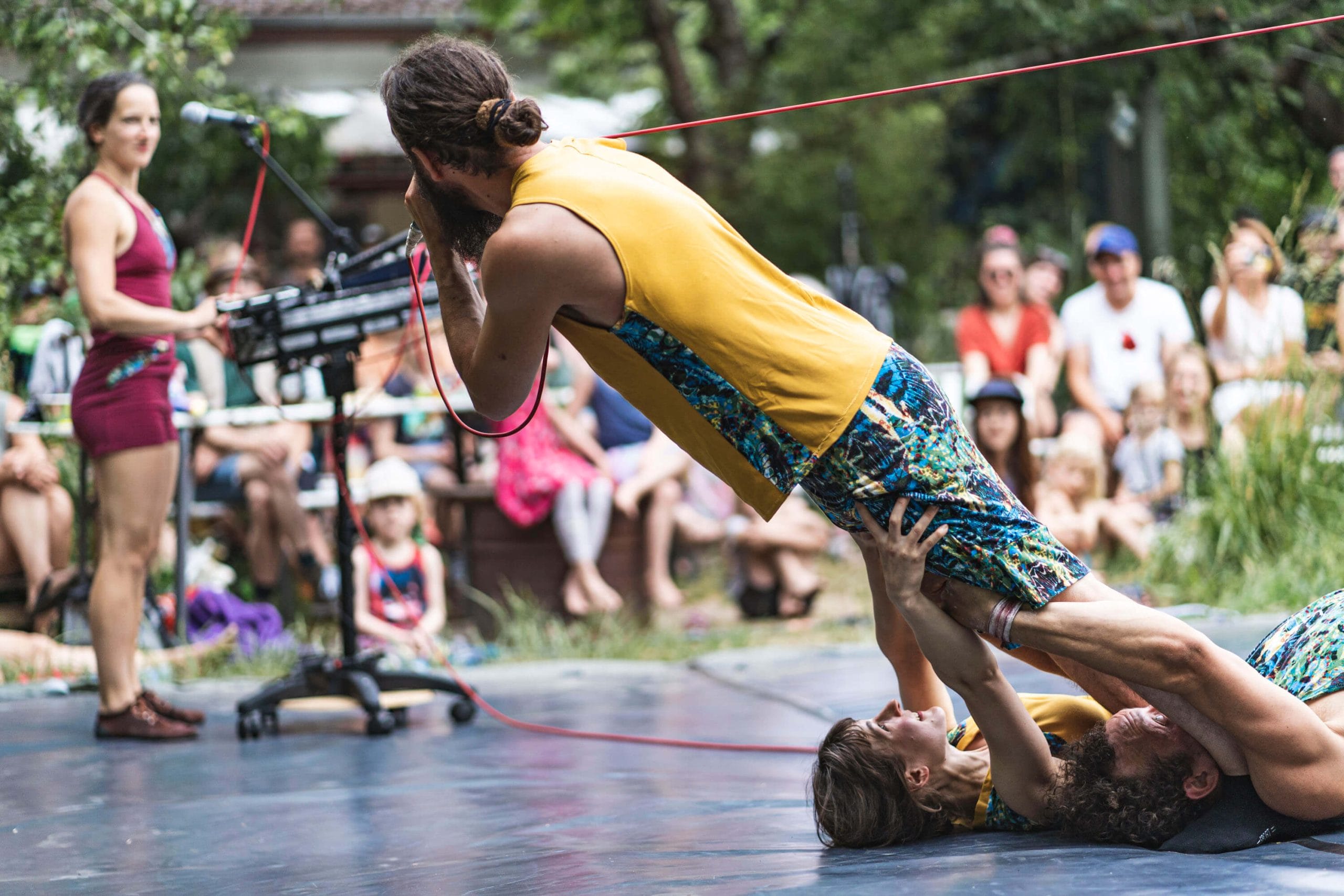 Two acrobats in colourful shorts and t-shirts lie on the floor, in front of an outdoor audience with their arms outstretched, holding onto a third acrobat who leaning forward held by a rope, a fourth performers stands in the background by a musical keyboard