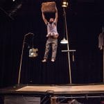 An acrobat in grey trousers and shirt with a dark beard, bounces on a trampoline holding a suitcase in the air.