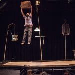 An acrobat in grey trousers and shirt with a dark beard, bounces on a trampoline holding a suitcase in the air.