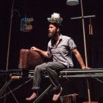 A man sits in the edge of trampoline holding a suitcase with a birds nest of his head.