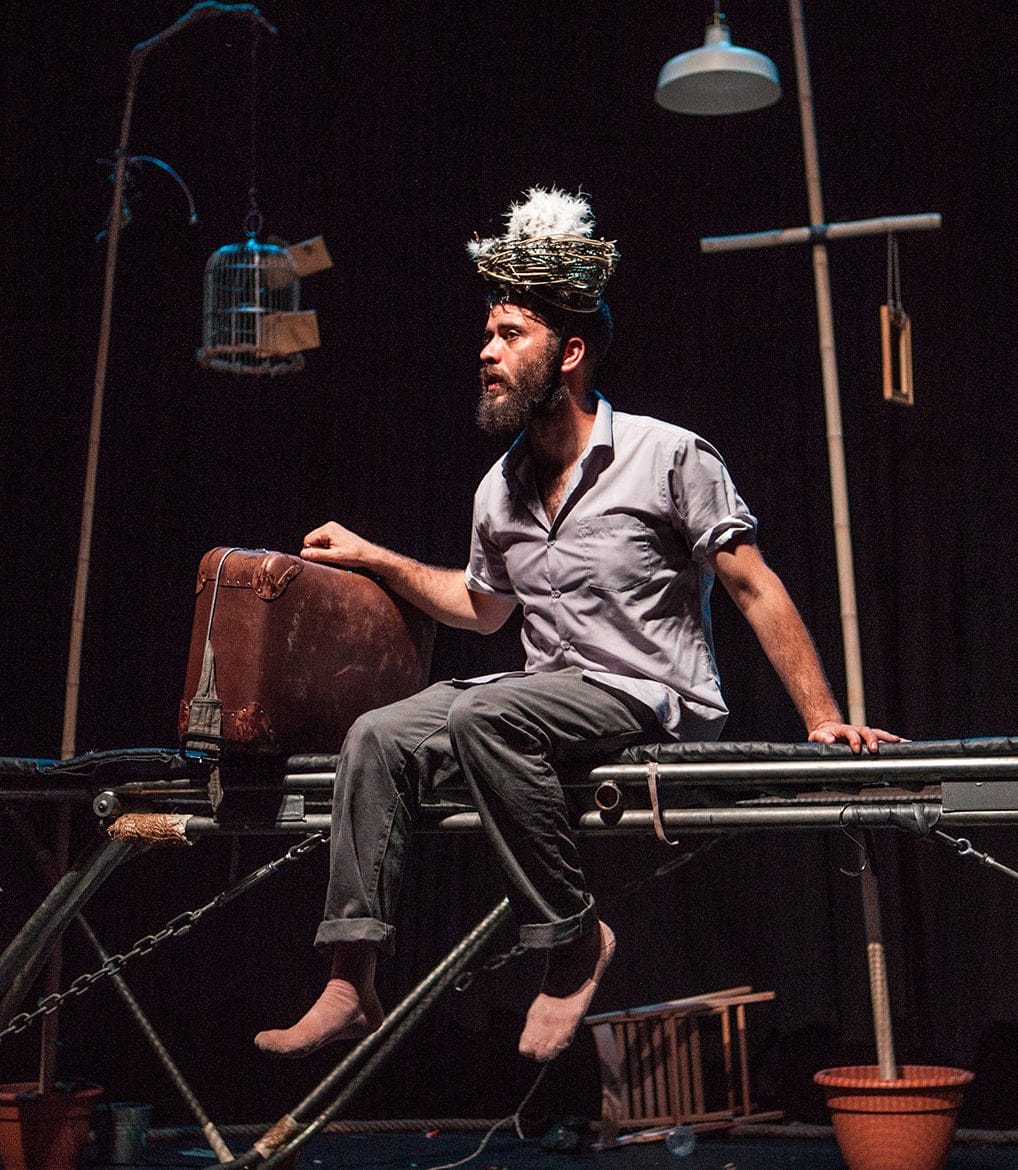 A man sits in the edge of trampoline holding a suitcase with a birds nest of his head.