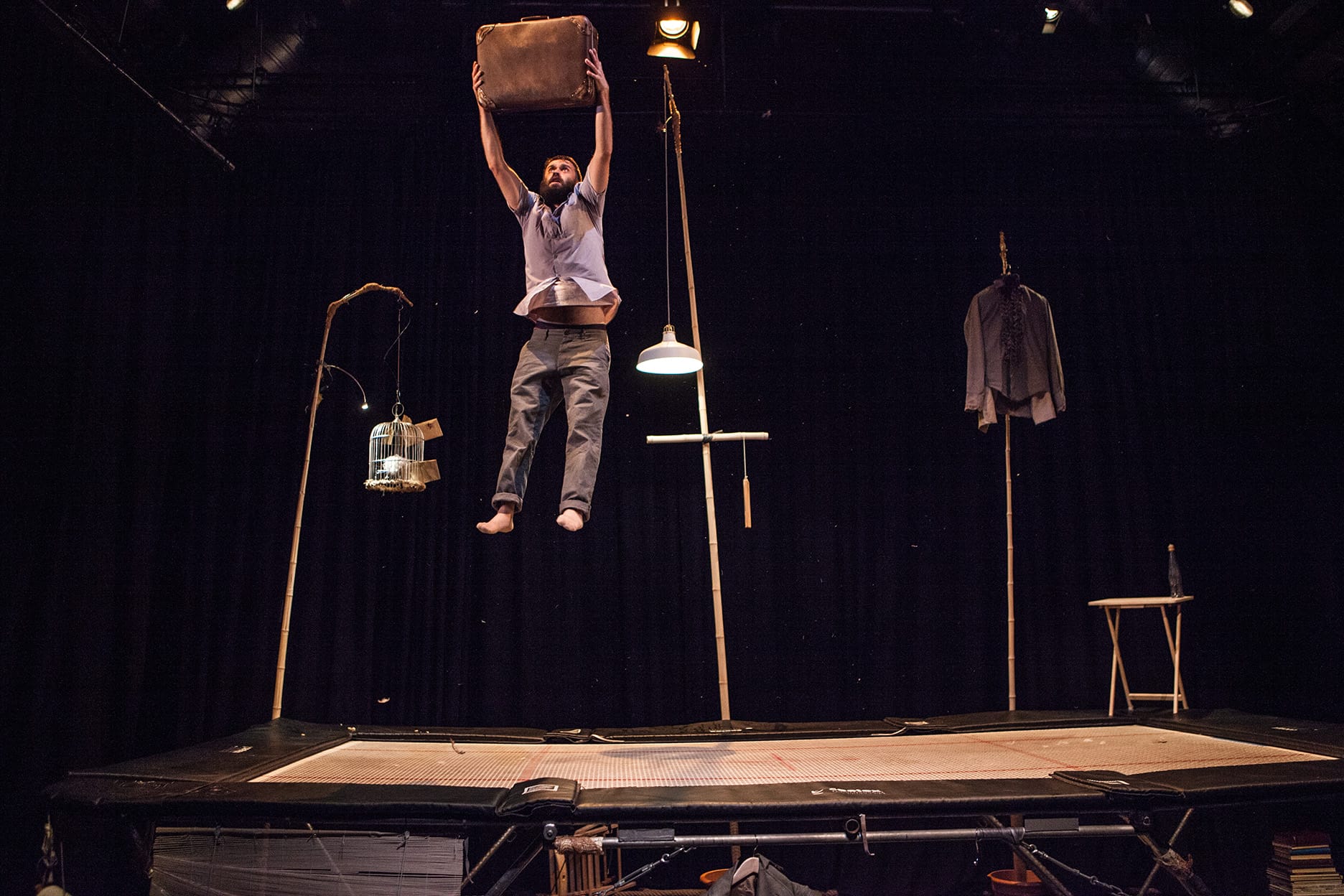 An acrobat in grey trousers and shirt with a dark beard, bounces on a trampoline holding a suitcase in the air.