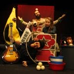 Four quirky performers are on stage, with one in a monkey mask lying down in front of the others while holding a guitar.