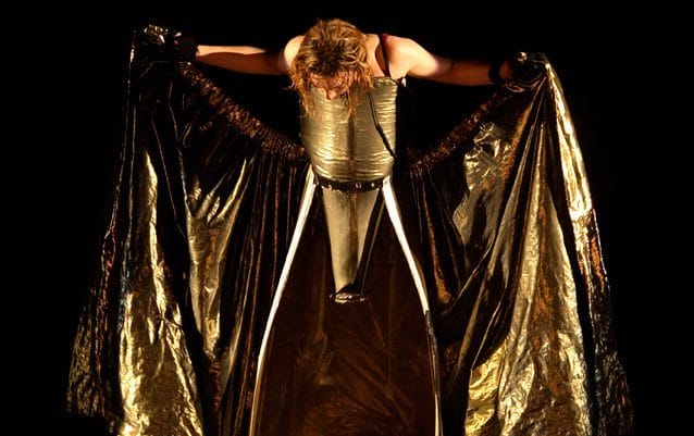 Performer with long blonde hair holds their outstretched golden wraparound skirt open at the front revealing an illusion of no legs, they are illuminated by a spotlight on a dark stage.