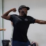 A performer dances in a pose as if holding a bow an arrow but with clenched fists, they wear a black t-shirt and black baseball hat and have dark facial hair.