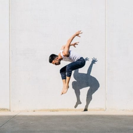 A person mid-air performing a dynamic jump in front of a sunlit wall, their shadow reflected on the surface.
