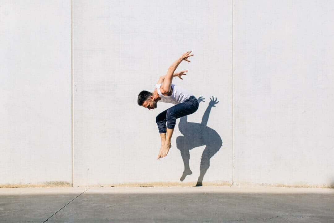 A person mid-air performing a dynamic jump in front of a sunlit wall, their shadow reflected on the surface.