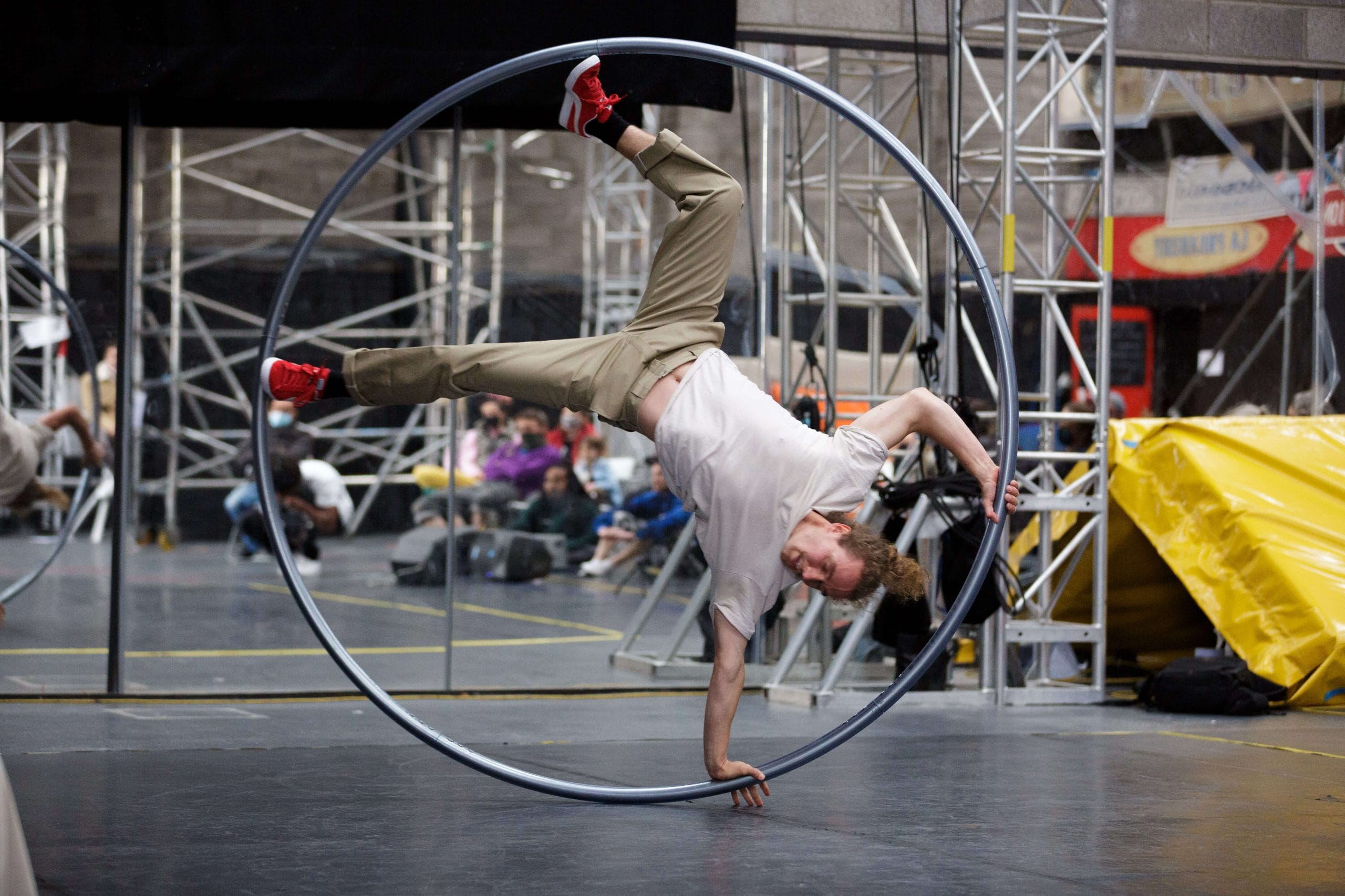 Performer rehearsing a handstand inside a large cyr whee;, with an audience in the background visible through a mirror's reflection.