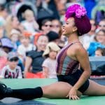 Performer in a black, purple and grey leotard and a pink flowered hairwrap and wearing roller skates, does the splits on stage, with an audience visible in the background.