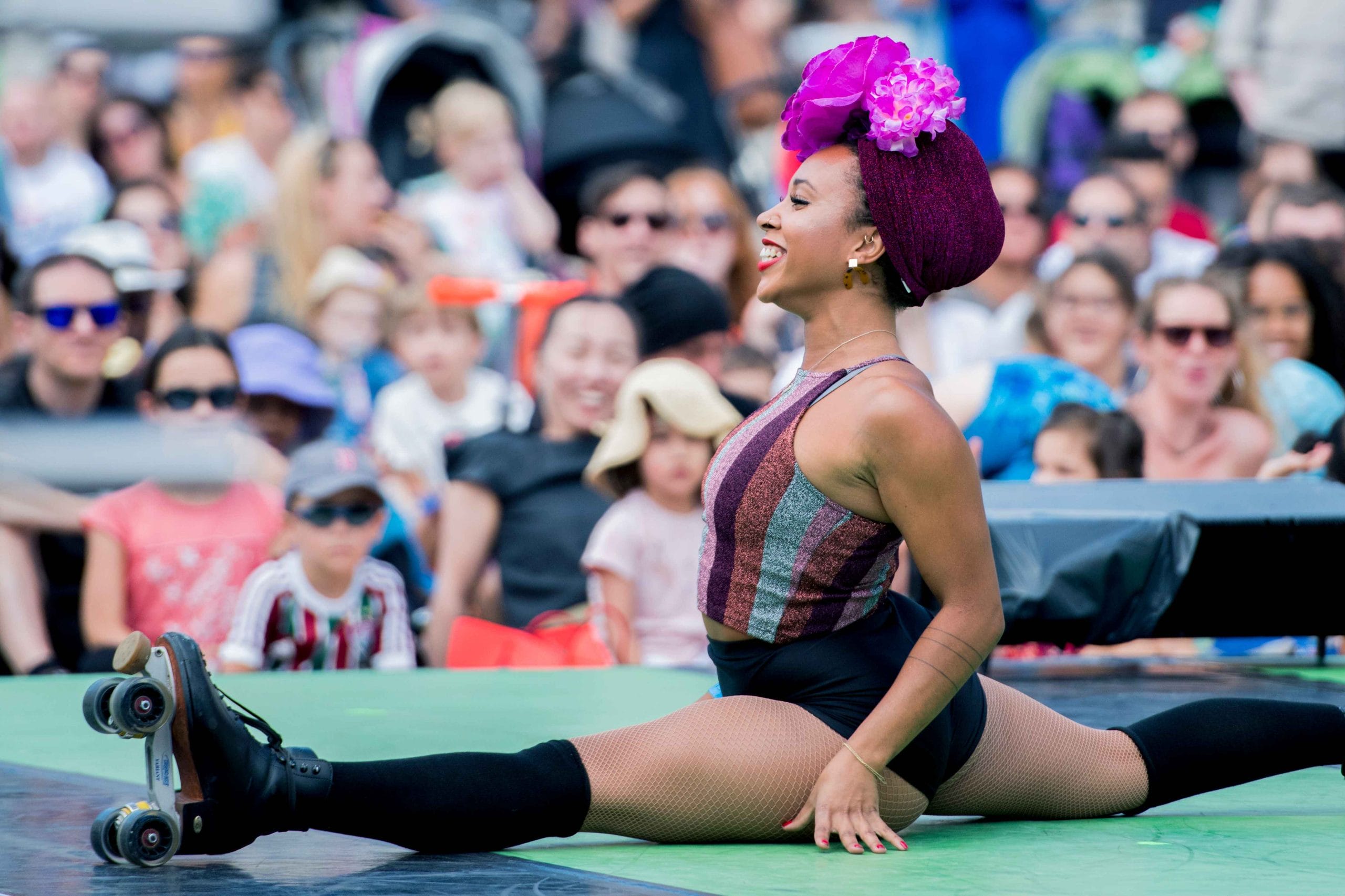 Performer in a black, purple and grey leotard and a pink flowered hairwrap and wearing roller skates, does the splits on stage, with an audience visible in the background.