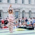 A performer in a shimmering tasseled pink outfit twirling multiple hula hoops at an outdoor event with an audience in the background.