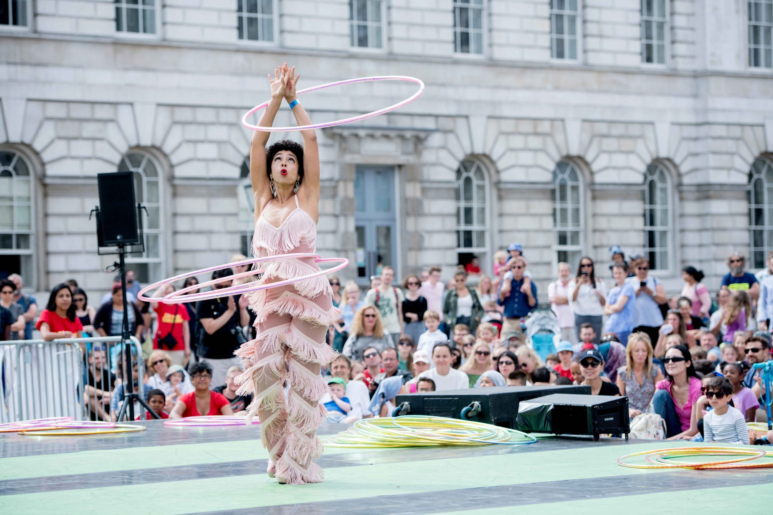 A performer in a shimmering tasseled pink outfit twirling multiple hula hoops at an outdoor event with an audience in the background.