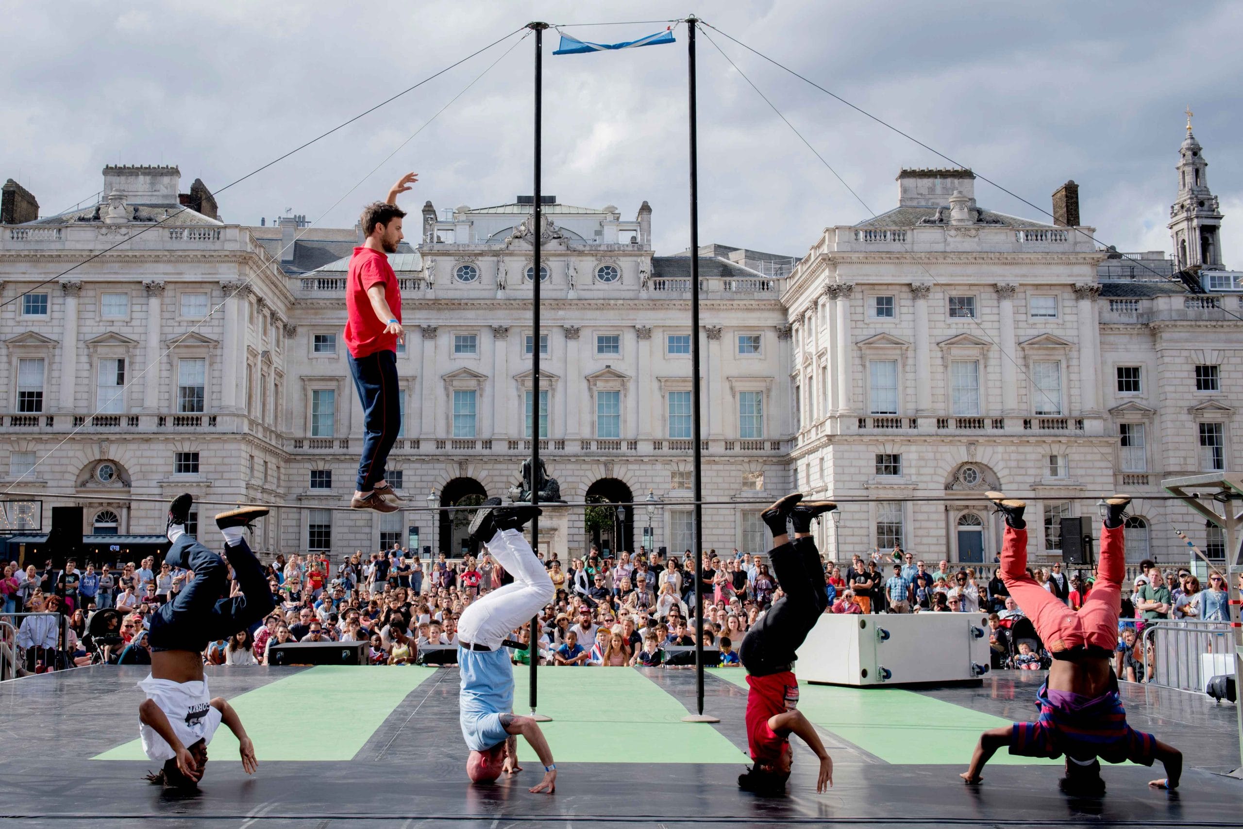 A tightrope walker and breakdancers perform before a crowd in front of a historic building.