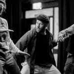 three performers stand in a row in a black and white indoor scene, their arms are outstretched towards each other creating movement
