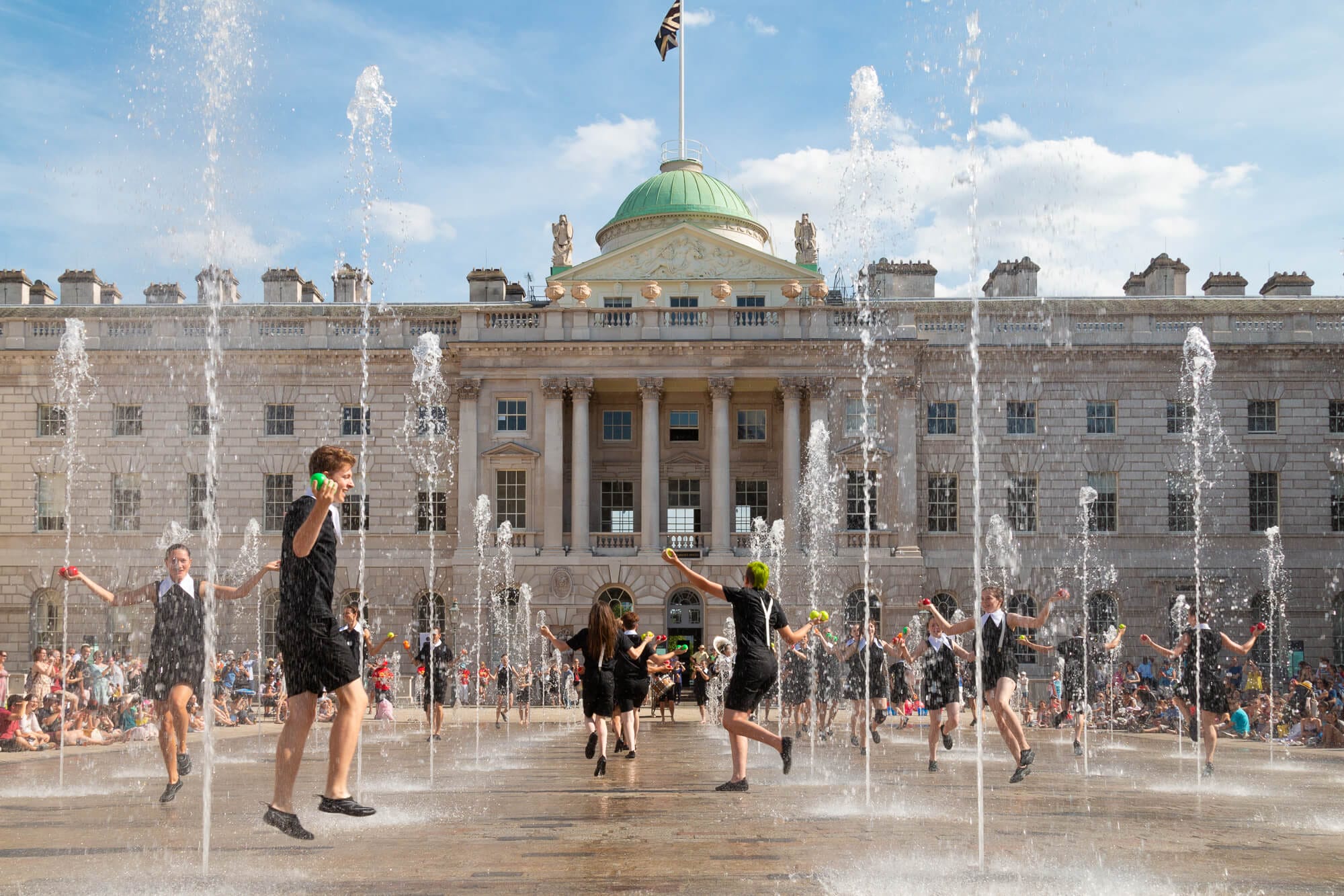 Several jugglers are dancing among fountain jets in an outdoor courtyard in front of a historic building with a flag on the roof with a crowd watching in the background, the sky is blue with a few bright white clouds