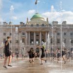 Performers juggling and dancing among fountains in front of a historic building, with a crowd watching in the background.