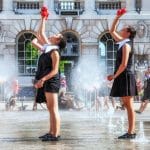 Two juggling performers part of Gandini Juggling, in coordinated black and white costumes juggle red balls among outdoor fountains in front of Somerset House in London. Spectators watch from the background.