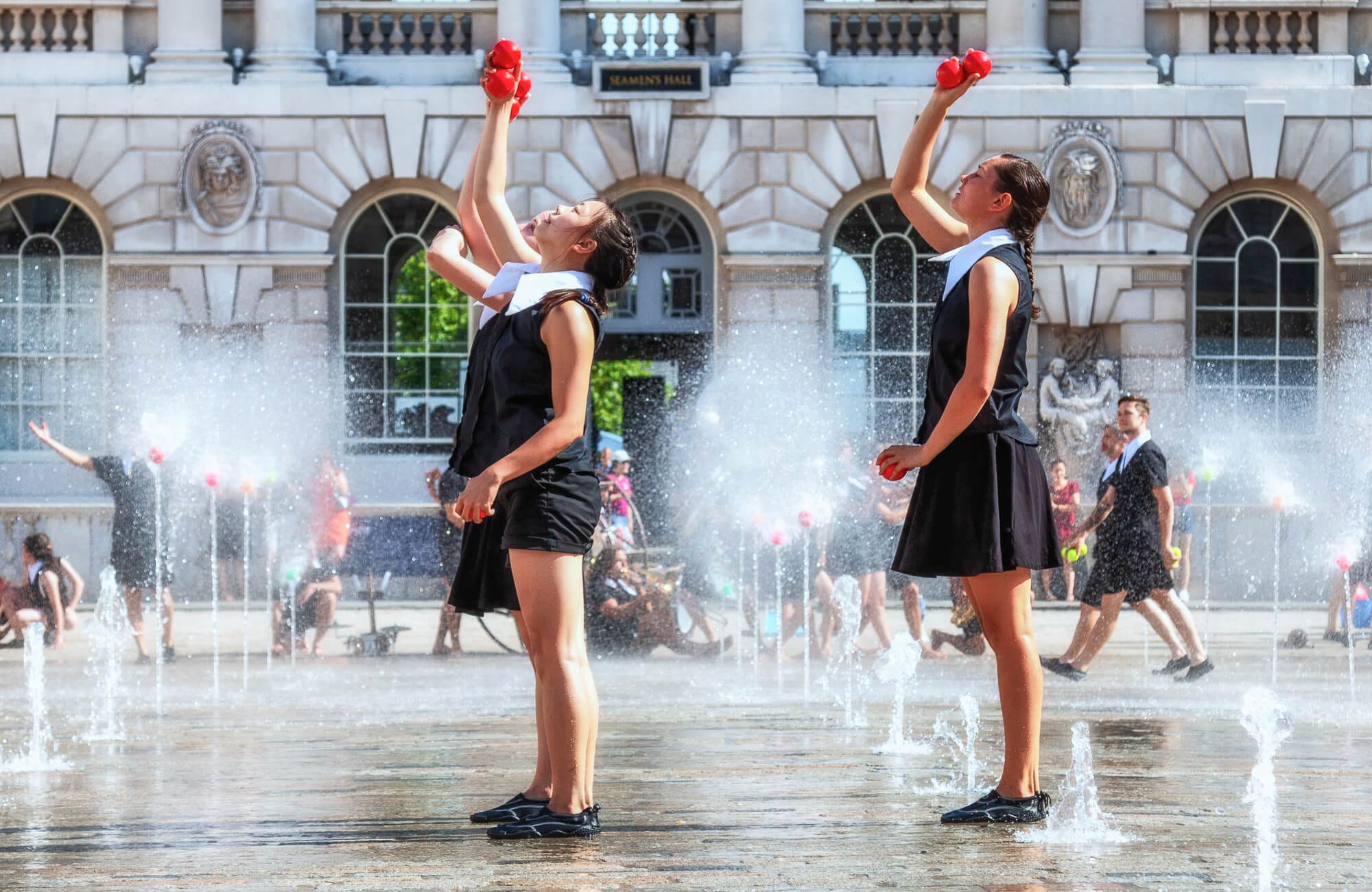 Two juggling performers part of Gandini Juggling, in coordinated black and white costumes juggle red balls among outdoor fountains in front of Somerset House in London. Spectators watch from the background.