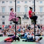Two performers on poles and four others lying on the stage floor, captivating a large outdoor audience in front of a historic building.