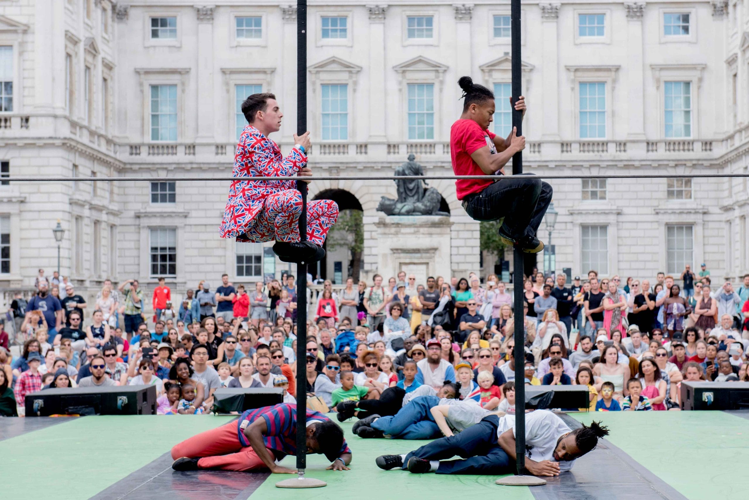 Two performers on poles and four others lying on the stage floor, captivating a large outdoor audience in front of a historic building.