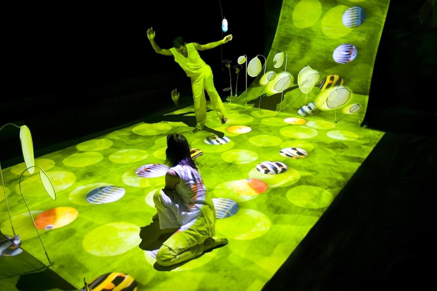 One performer sits while the other strikes an artistic pose, standing on a green-illuminated stage.