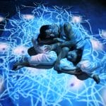 The two performers lie closely intertwined on their sides on the blue-toned stage.