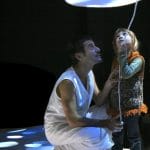 A theatrical performance featuring a performer kneeling next to a child, as they together hold a mushroom-shaped lamp above them, casting a soft, warm glow.