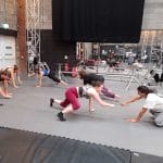 Group of people practicing acrobatics in a gym, with stage equipment visible in the background.