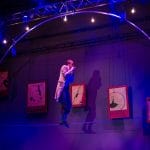 a tightrope walker in a long coat walks delicately across the rope, whilst playing the mouth organ, their shadow is created on the wall behind by a purple coloured spotlight