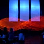 Five children sit on the floor in a darkened room watching a light display in front of them with bright blue and orangey red lights